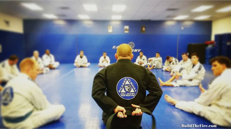 An instructor in a black gi faces students seated around the mat
